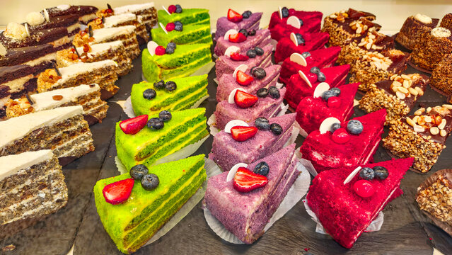 Rows of colorful cake slices sit on a display tray in a bakery shop. The assortment includes vibrant fruit-topped layers, nut-covered pieces, and creamy dessert sponges