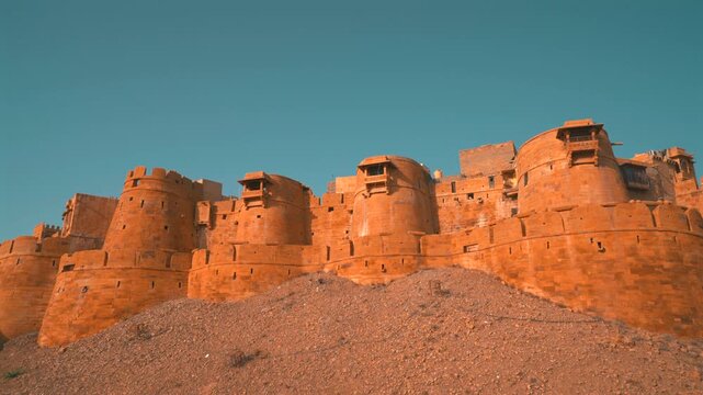 Cinematic low angle static shot of the massive golden sandstone bastion walls of Jaisalmer Fort standing on a rocky hill under a blue sky in Rajasthan, India.