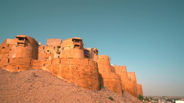 Cinematic low angle static shot of the imposing golden sandstone bastions of Jaisalmer Fort standing above a gravel hill under a clear sky in Rajasthan, India.