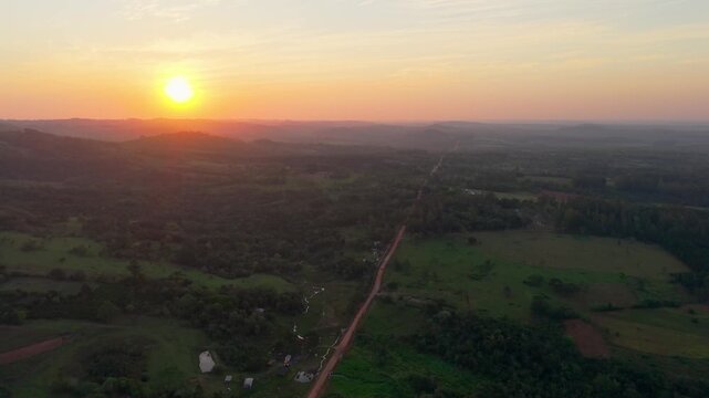 Drone video captures a golden sunrise illuminating rolling hills, lush greenery, and a rural road weaving through the vast landscapes of Nueva Alborada under soft pastel skies.