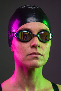 Gros plan du visage d'une nageuse de comp&eacute;tition, qui porte des lunettes de piscine noires opaques, un bonnet de bain en caoutchouc noir. Expression neutre et &eacute;clairage rose et vert - vertical
