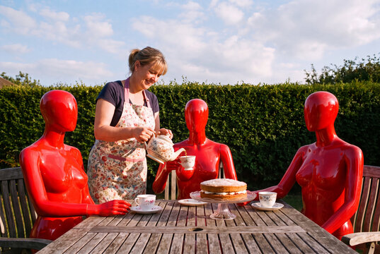 Une femme de 50 ans avec un tablier de cuisine sert le th&eacute; sur sa terrasse &agrave; 3 mannequins de vitrine rouge par beau temps - surr&eacute;alisme - horizontal