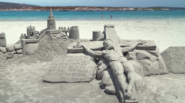 Sand sculpture of Jesus on the cross with John 3:16 plaque and castle on the beach