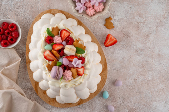 Easter egg-shaped Pavlova cake with whipped cream, topped with fresh berries and assorted sweets on a wooden board on a concrete background. Top view, copy space.