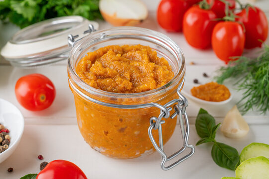 Zucchini caviar in a glass jar with fresh vegetables and herbs on a white wooden background, homemade vegetable spread for healthy eating and canning.