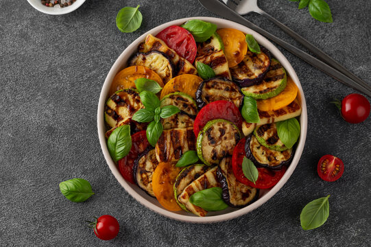 Grilled vegetable salad with zucchini, eggplant, tomatoes, halloumi cheese, basil and balsamic glaze on a plate on a dark concrete background. Healthy food. Top view.