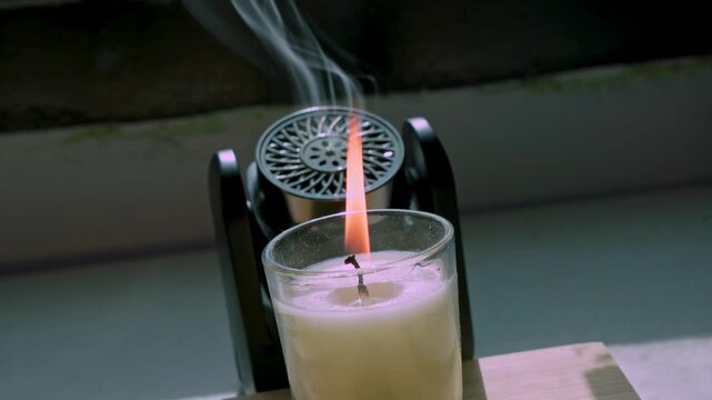 Close-up of a burning white candle in a glass jar next to a black aroma diffuser emitting a thin mist. The objects rest on a wooden surface indoors, illuminated by bright natural daylight.