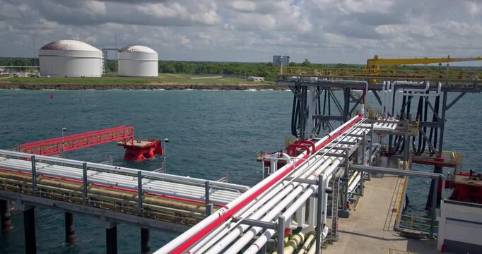 Static high-angle wide shot showing fuel transport pipelines along the jetty towards the marine unloading platform and spherical LPG storage tanks on the coastline at Coastal Petroleum terminal in San