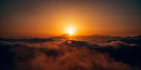 Obraz premium A sunrise over a mountain range with clouds and mist glowing in warm golden light. Dramatic sky and serene landscape, a peaceful and inspiring natural scene