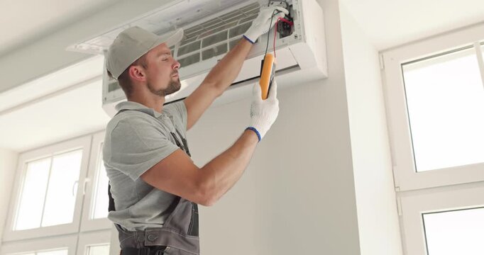 HVAC technician performs airconditioner testing with multimeter. Worker checks wiring inside wall mounted cooling unit in bright home. Reliable climate control service for modern comfort.