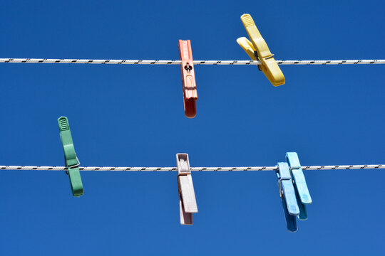 Five vibrant plastic clothespins in various colors hang on two white washing lines against a clear blue sky background.