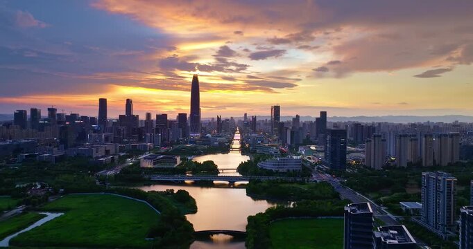 Aerial view of a modern skyline at sunset with a supertall skyscraper, waterways, bridges, and lush urban parks beneath colorful evening clouds.
