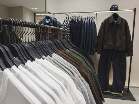 Men's clothing displayed on retail racks, featuring white shirts, gray tops, dark trousers, brown jacket, showcasing organized fashion assortment, modern store interior for apparel merchandising