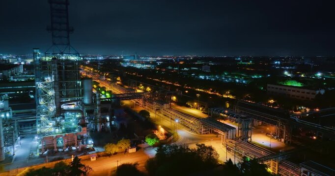 High angle night view of an illuminated chemical plant and oil refinery with storage tanks, industrial pipelines, steel structures, and lights.