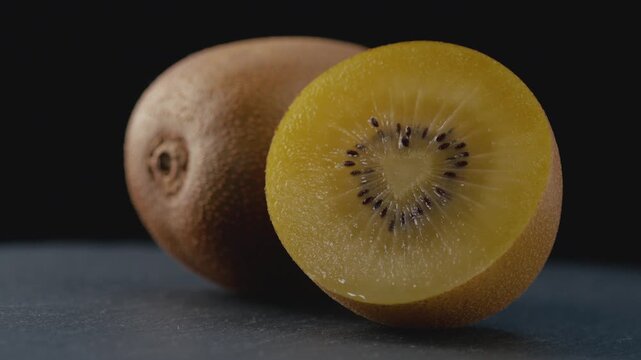 Macro shot of a ripe golden kiwi cut in half with a heart-shaped core, rich juicy pulp and seeds on a dark background with a blurred whole kiwi for healthy eating, gourmet food, and culinary content.