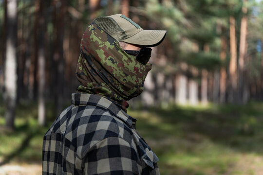 Side view of tactical instructor wearing camouflage balaclava dark glasses and cap standing in pine forest environment during outdoor training session