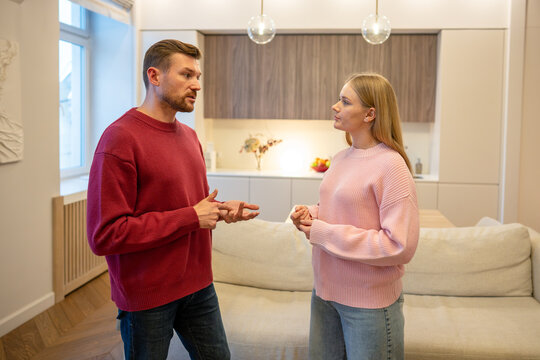 Positive married couple man and woman standing in living room at home engaged in conversation, male partner explaining and sharing thoughts with loving female, family relationships and partnership.