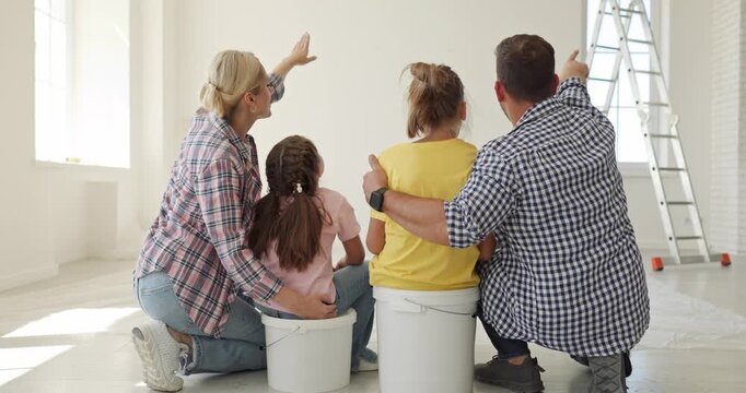 Parents children plan home renovation together. Rear view of family in an empty room, pointing at a wall beside a ladder and imagining future interior. A hopeful vision of family life ahead.