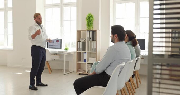 Business speaker leads training for colleagues. Male professional presents chart data to seated coworkers during an office meeting in a workplace. Corporate teamwork supports professional growth.