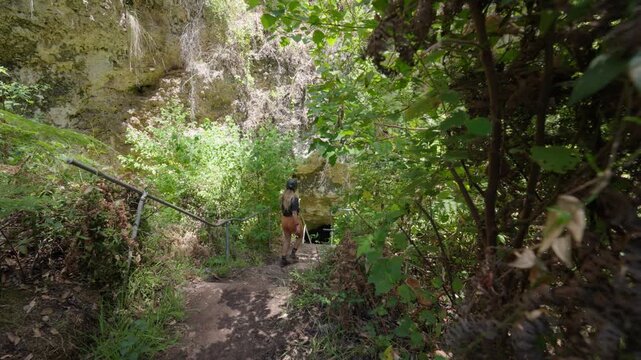 A woman carefully making her way down a lush forested path and into the entrance pf a mysterious cave surrounded by dense green vegetation and rugged natural rock formations