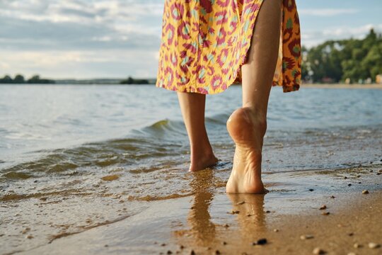 Unrecognizable woman strolls barefoot on a sandy beach, waves gently lapping at her feet. Vibrant colors of her dress contrast with the calm waters as the sun sets in the background, creating a serene atmosphere