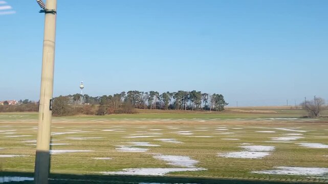 Open snow patched field under blue sky, distant tree line and tower, scattered thawing snow on grass, utility pole and wires. View from train window. Video taken on mobile device.