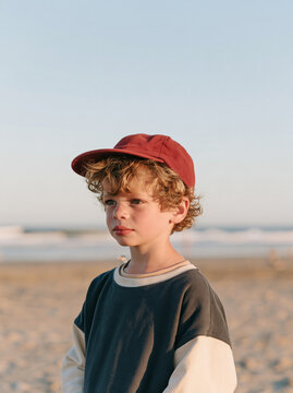Portrait of young boy wearing red baseball cap on beach with ocean in background