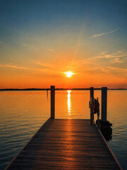 Fototapeta premium Warm sunset reflects across calm water beside a simple wooden pier, creating a peaceful minimalist seascape with glowing sunlight, soft colors, silhouettes, and tranquil evening atmosphere.