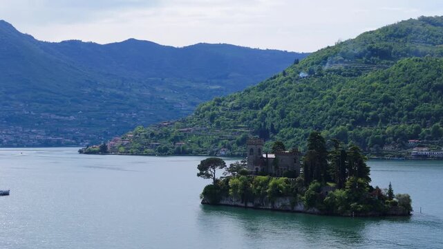 A cinematic aerial shot of the historic castle on Isola di Loreto in Lake Iseo, Italy, surrounded by lush mountains and calm blue water.