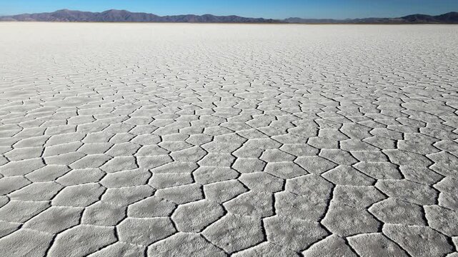 Wide view of Salinas Grandes salt flat with dry polygon patterns stretching toward distant mountains under clear blue sky, useful as abstract landscape or climate change b-roll.