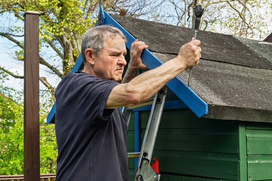 Older man standing on stepladder uses screwdriver to remove timber fascia board from pitched felt roof of small painted wooden garden shed or playhouse during DIY maintenance
