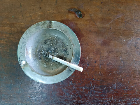 A classic metal ashtray with a lit cigarette butt sits on an old wooden table.