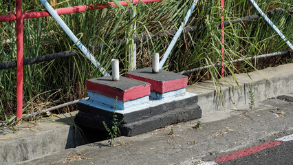 Twin concrete flag pole stands painted red, blue, and black placed along an outdoor concrete pathway and drain. © agung