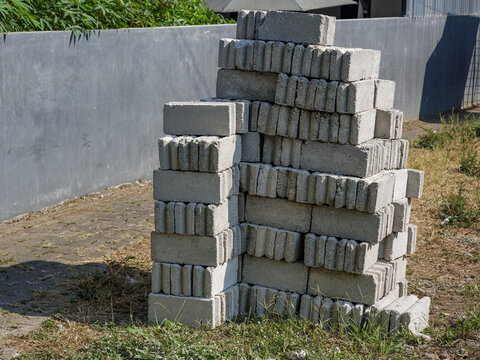 Grey concrete cinder blocks stacked together on ground near a concrete boundary fence in sunlight.