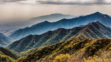 Obraz premium Layers of blue mountain ridges fade into distant haze under cloudy sky. Green forested slopes show autumn colors. Remote valley below is partly visible.