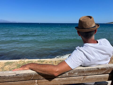 Uomo con cappello di paglia seduto su una panchina mentre guarda il mare blu in estate. Artemis, Grecia