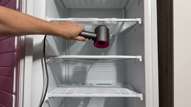 Man defrosts a freezer using a hairdryer's hot air, ice chunks falling and melting, puddles on floor, with towels and bucket, candid authentic life domestic maintenance task at home.