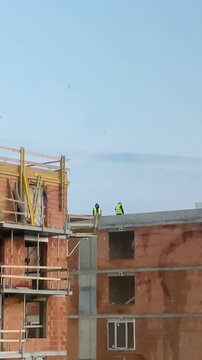Roofers on rooftop inspecting parapet and wearing highvisibility vests while checking scaffolding and brickwork against clear blue sky, slow deliberate motions. Vertical shot taken on mobile device
