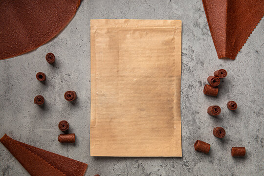 Natural fruit leather rolls and sheets arranged around craft paper ziplock pouch mock up on grey concrete background