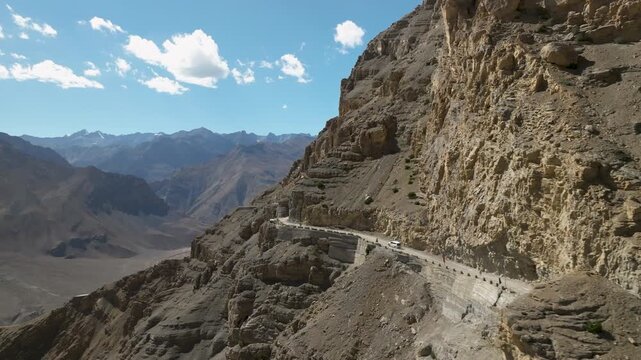 Aerial video of a vehicle driving on a dramatic cliffside road carved into a towering mountain face overlooking a vast valley under a blue sky in Spiti Valley Himachal Pradesh India shot by a drone