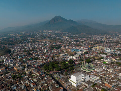 Aerial view of Batu city with Mount Panderman in the background, featuring dense urban rooftops, a prominent mosque, and a Ferris wheel in Batu, East Java, Indonesia.