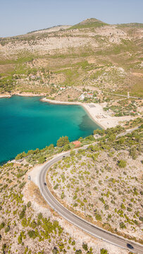 Aerial view of a coastal road winding along the hillside above a turquoise bay with a sandy beach and Mediterranean scrubland, Thasos, Greece.