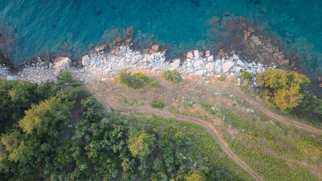 Aerial view of the rocky coastline with turquoise water, lush green trees, and a dirt path along the shore in Thasos, Greece.