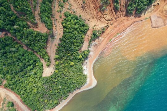 Aerial view of PT Ceria Nugraha Indotama mining site showing red soil erosion and lush forest meeting the turquoise sea in Wolo, South East Sulawesi, Indonesia.
