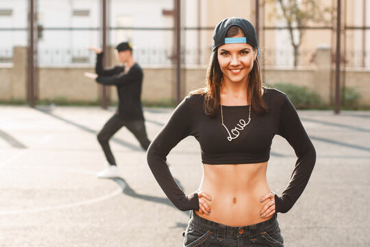 Confident young woman in urban streetwear (crop top, backward cap, 'Love' necklace) poses confidently on an outdoor basketball court. Blurred man dances in background. Bright sun, golden hour light.