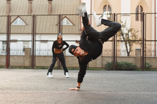Athletic male breakdancer performing a dynamic one-hand freeze trick on an outdoor court as a stylish young woman watches in the background. Hip-hop youth culture, street dance, and active lifestyle.