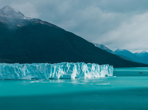 Aerial view of Perito Moreno Glacier with its ice wall above turquoise waters, set against dark mountains under a cloudy sky in Perito Moreno, Santa Cruz Province, Argentina.