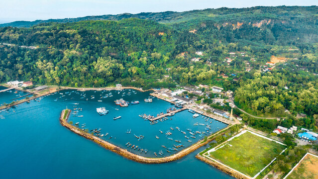 Aerial view of Dermaga Tamperan fishing port with boats anchored in the blue bay, surrounded by lush green hills and coastal vegetation in Kecamatan Pacitan, Jawa Timur, Indonesia.