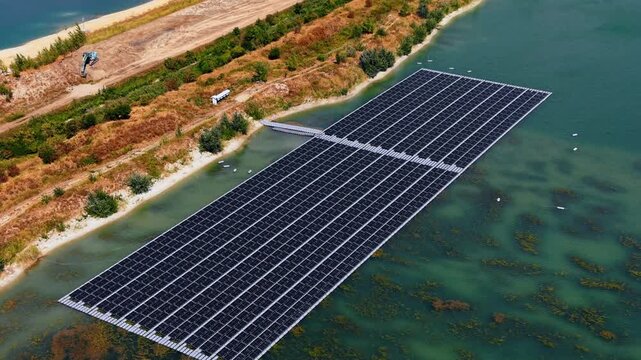 High angle view of photovoltaic panels on a freshwater reservoir. A rectangular pontoon system supporting energy generation technology floats near a sandy embankment covered with bushes.