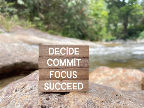 Inspirational wooden sign displaying decide, commit, focus and succeed on a rocky riverside, representing motivation, determination and personal goals.